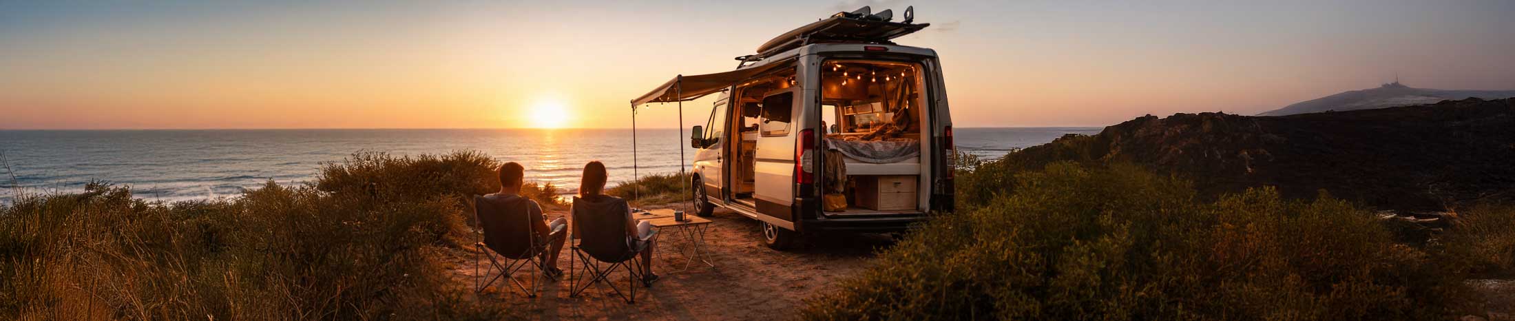 Paar sitzt vor einem ausgebauten Campervan und blickt bei Sonnenuntergang auf das Meer